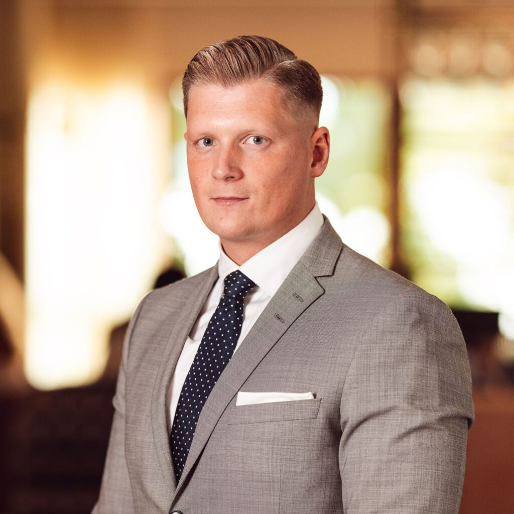 Headshot of Patrik Westræus in formal attire with a neatly combed hairstyle, looking directly at the camera. He is wearing a light grey suit with a white shirt and a dark blue tie with polka dots. A white pocket square complements his suit. The background is softly blurred with warm lighting, suggesting an upscale indoor setting.