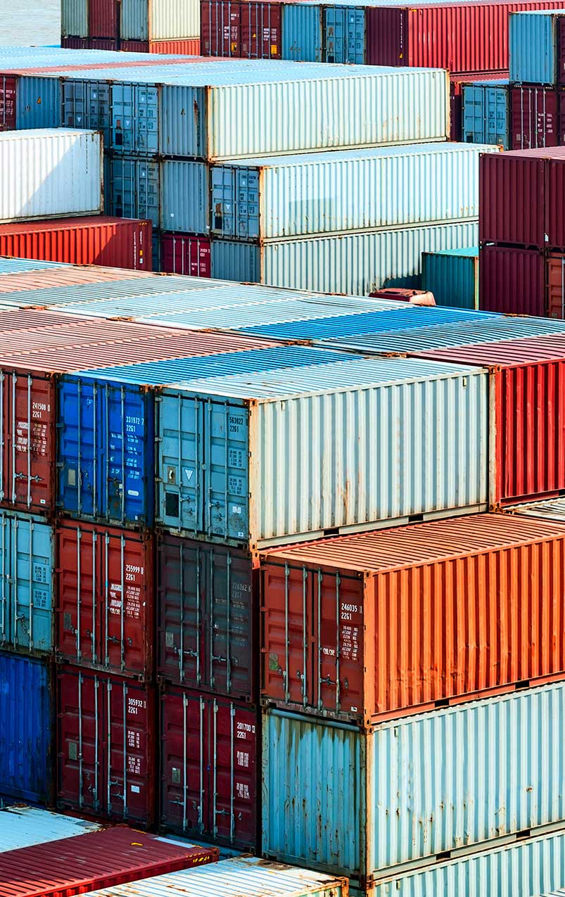 ALT Text: "Stacks of colorful shipping containers in a cargo port. The containers are arranged in an orderly fashion, showcasing a variety of colors such as red, blue, and white, with visible numbers and letters indicating their identification. The image captures the organized chaos typical of shipping yards.