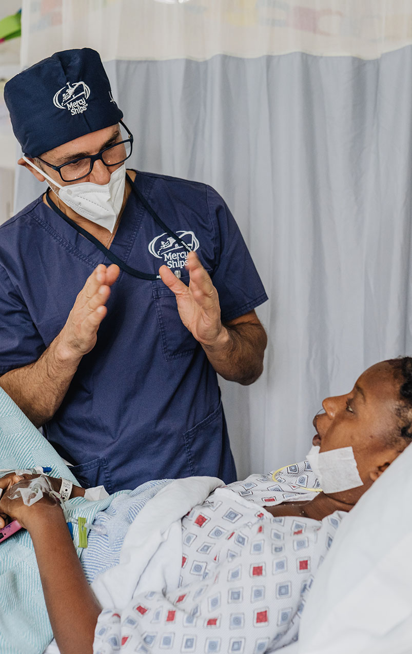 A medical professional wearing a scrub cap and mask with the "Mercy Ships" logo is standing beside a bed and talking to a patient. The patient is lying in bed, appearing attentive to the doctor, with a bandage on their neck and an IV line in their arm. The setting suggests a hospital room with a privacy curtain in the background.