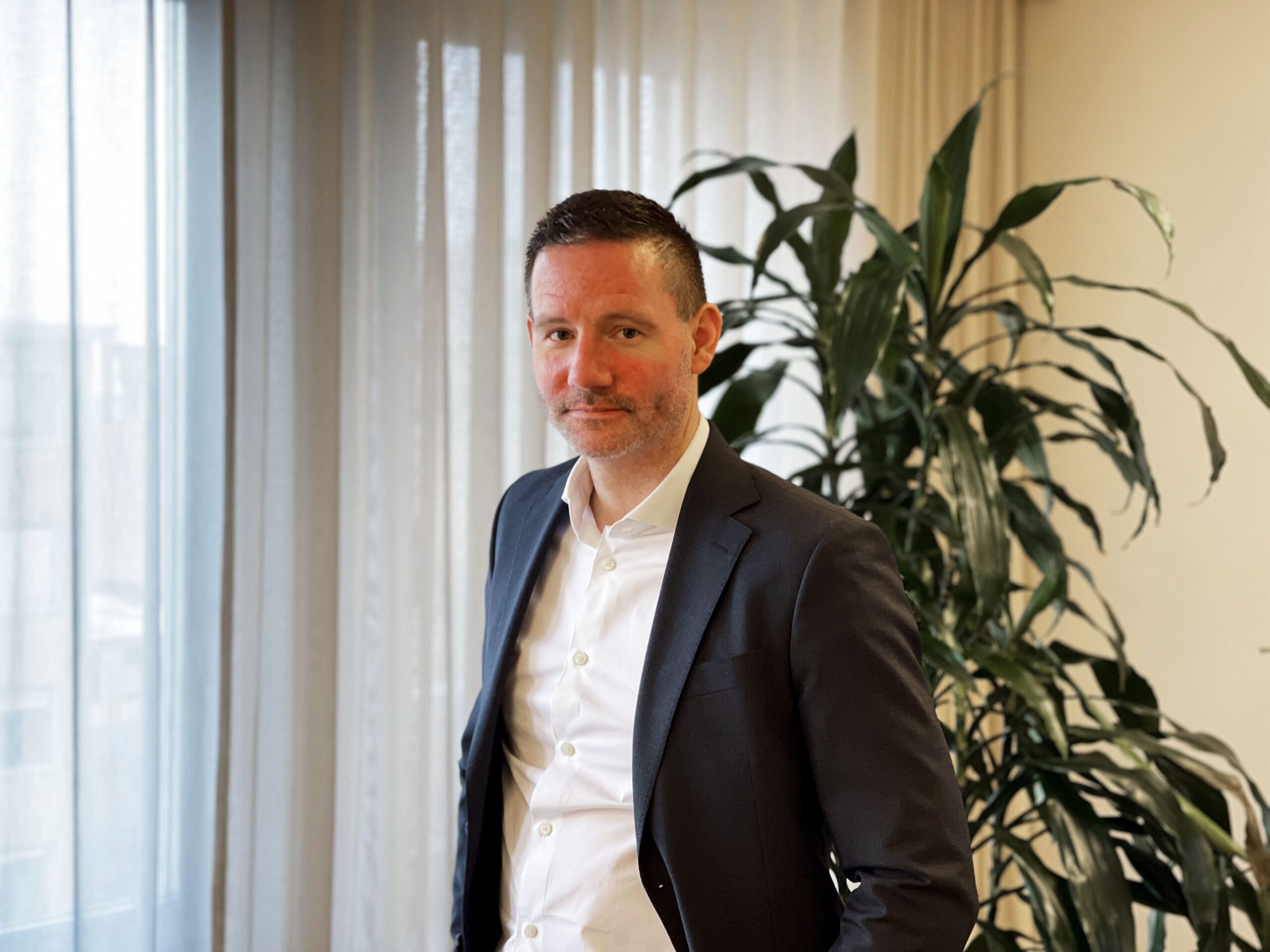 En man med kort mörkt hår och skäggstubb står leende mot kameran. Han är klädd i en mörk kostym med vit skjorta. Bakgrunden visar ett ljust kontorsutrymme med genomskinliga gardiner och en stor grön växt.