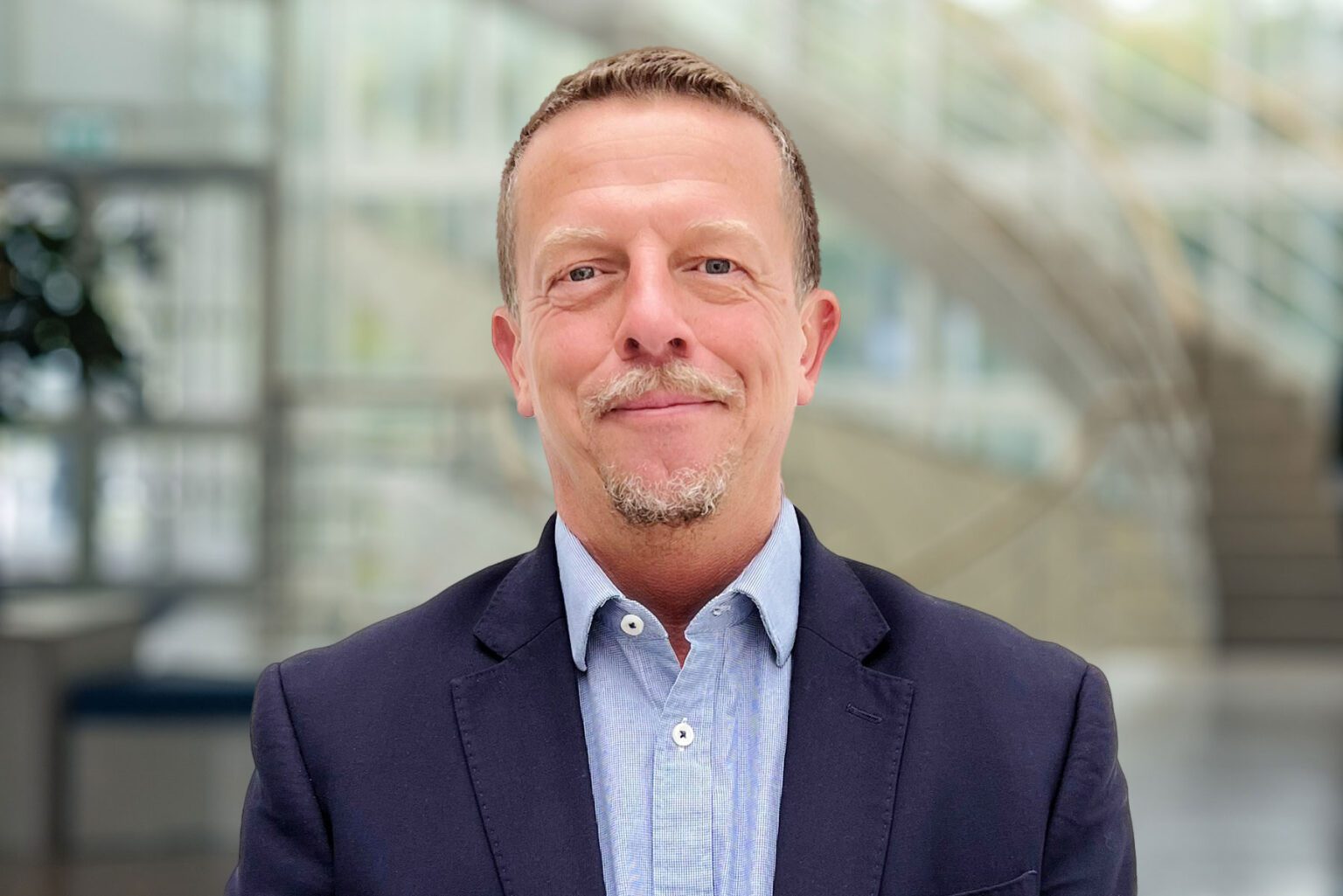 A headshot of a middle-aged man with a friendly demeanor, featuring short cropped hair and a goatee. He is wearing a navy blazer over a blue button-up shirt. The background is blurred with architectural details suggesting a modern office with abundant natural light.