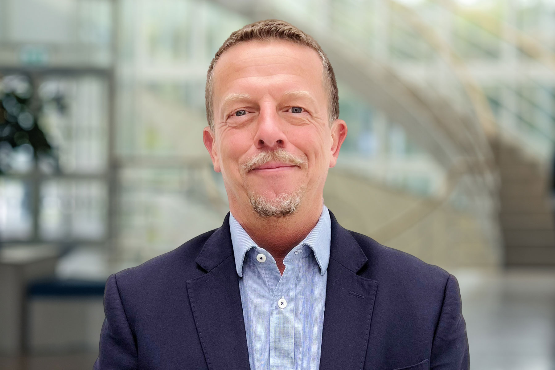 A headshot of a middle-aged man with a friendly demeanor, featuring short cropped hair and a goatee. He is wearing a navy blazer over a blue button-up shirt. The background is blurred with architectural details suggesting a modern office with abundant natural light.