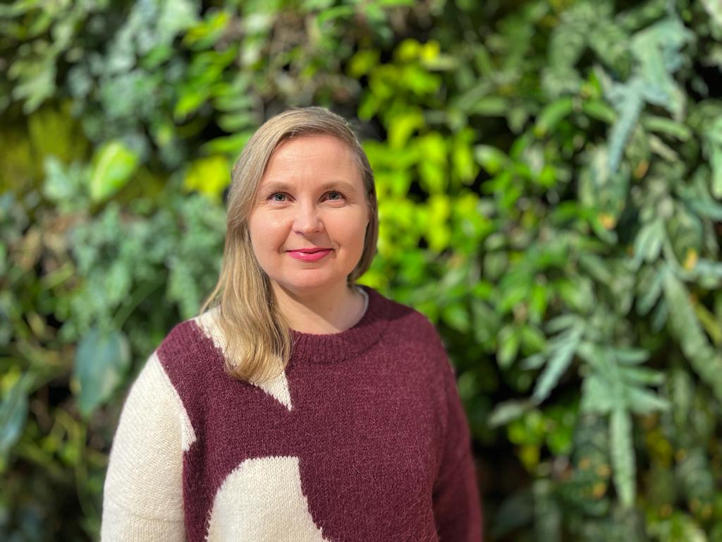 A cheerful woman with blonde hair, wearing a cream and burgundy patterned sweater, is smiling at the camera. The background features a lush green wall of plants with various shades of green leaves, creating a vibrant and natural backdrop.